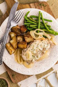white dinner plate containing one slow cooker pork chops with onions, green beans and potato wedges with dinner knife and fork on top of a wooden cutting board