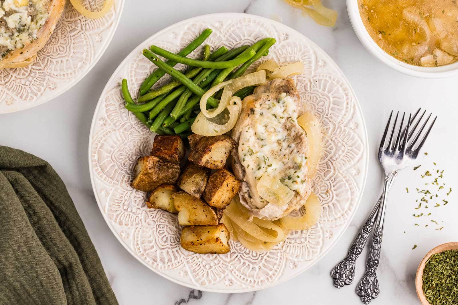 white dinner plate containing one slow cooker pork chops with onions, green beans and potato wedges with dinner forks on side