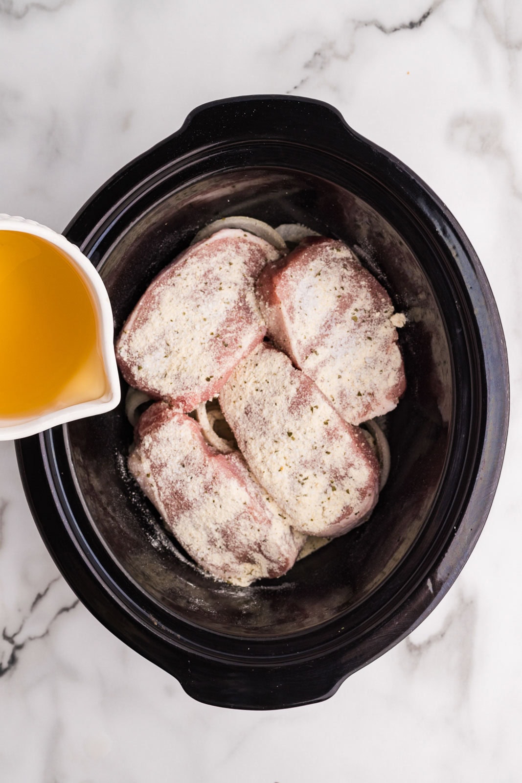 overhead view of sliced onions and pork chops that have been sprinkled with ranch seasoning and container of chicken broth being poured around edges