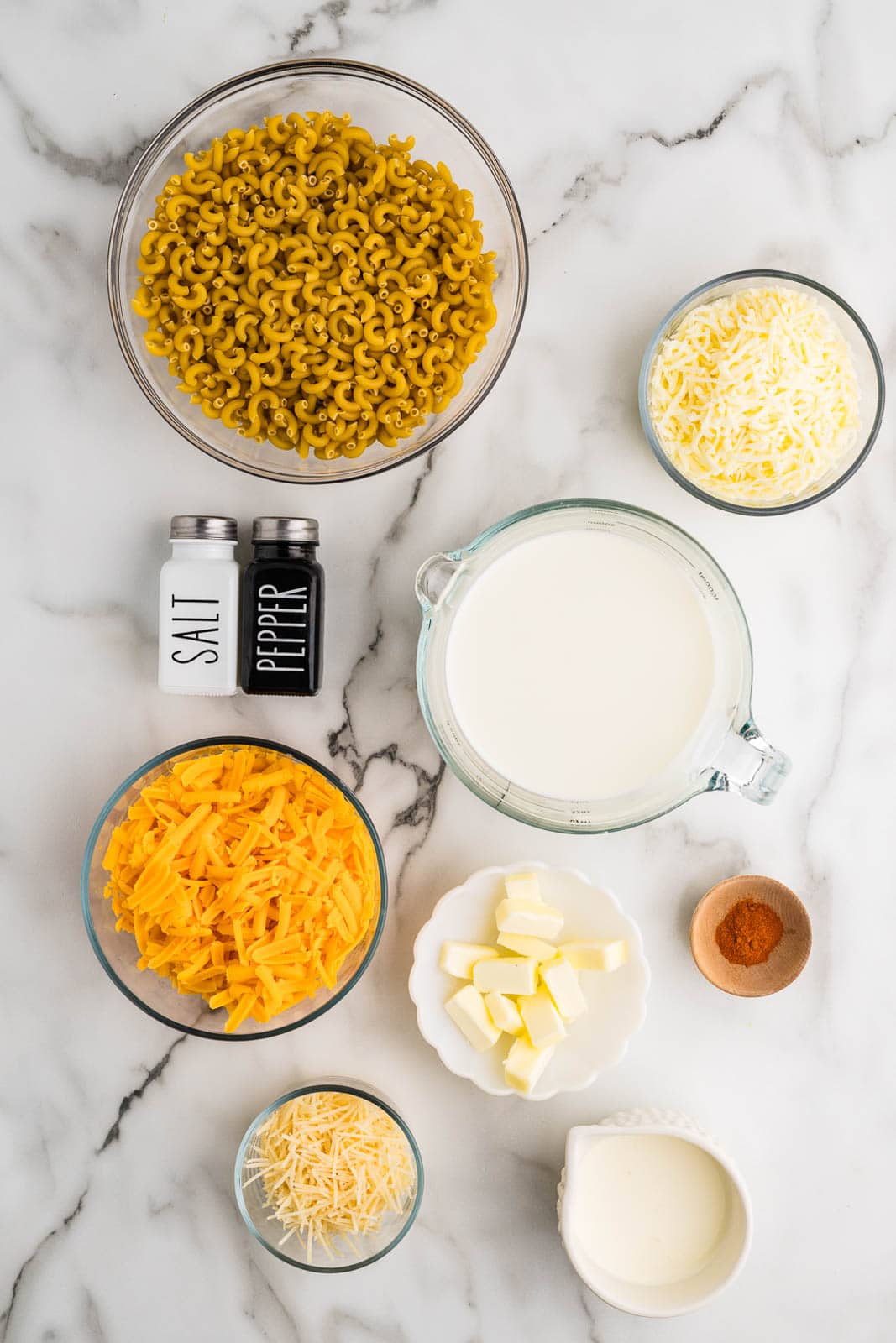 Mac and cheese ingredients to include bowls of elbow macaroni, shredded cheddar cheese, shredded Monterey Jack cheese, shredded Parmesan cheese, cut up butter, measuring cups of whole milk and cream, and and pepper shakers, and small bowl of smoked paprika