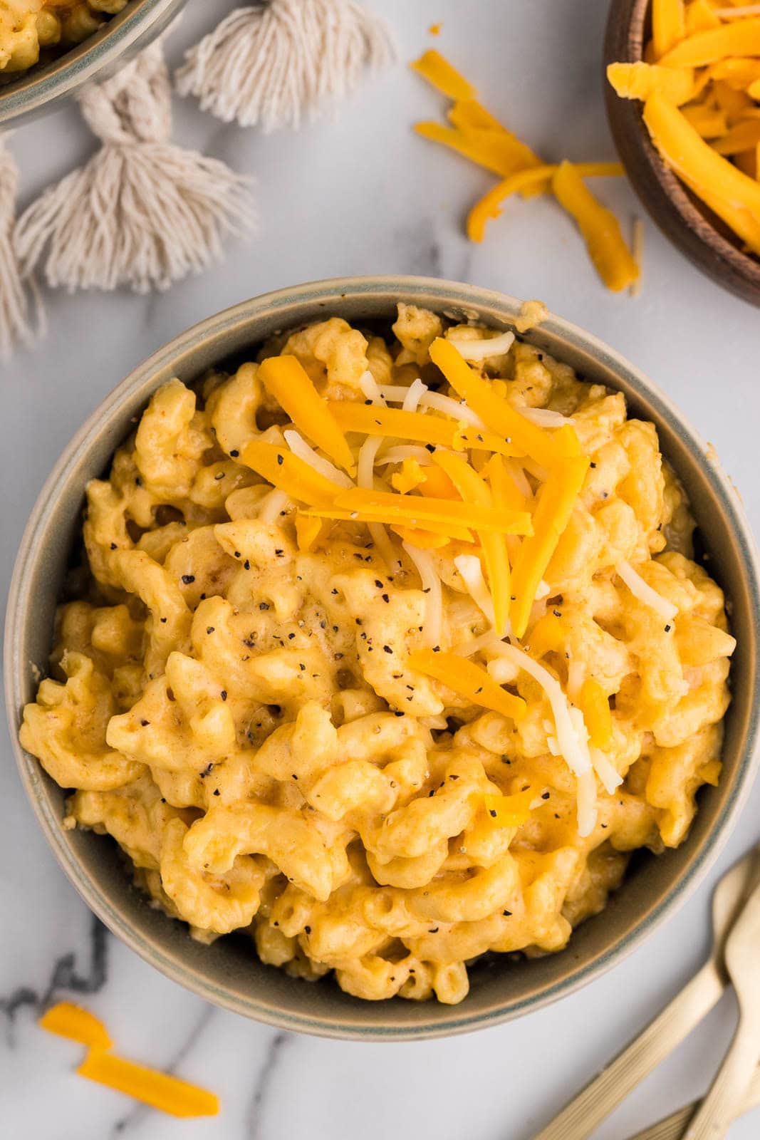 overhead view of bowl of Mac and cheese with a small amount of shredded cheese on top, surrounded by a bowl of cheese and forks on the side