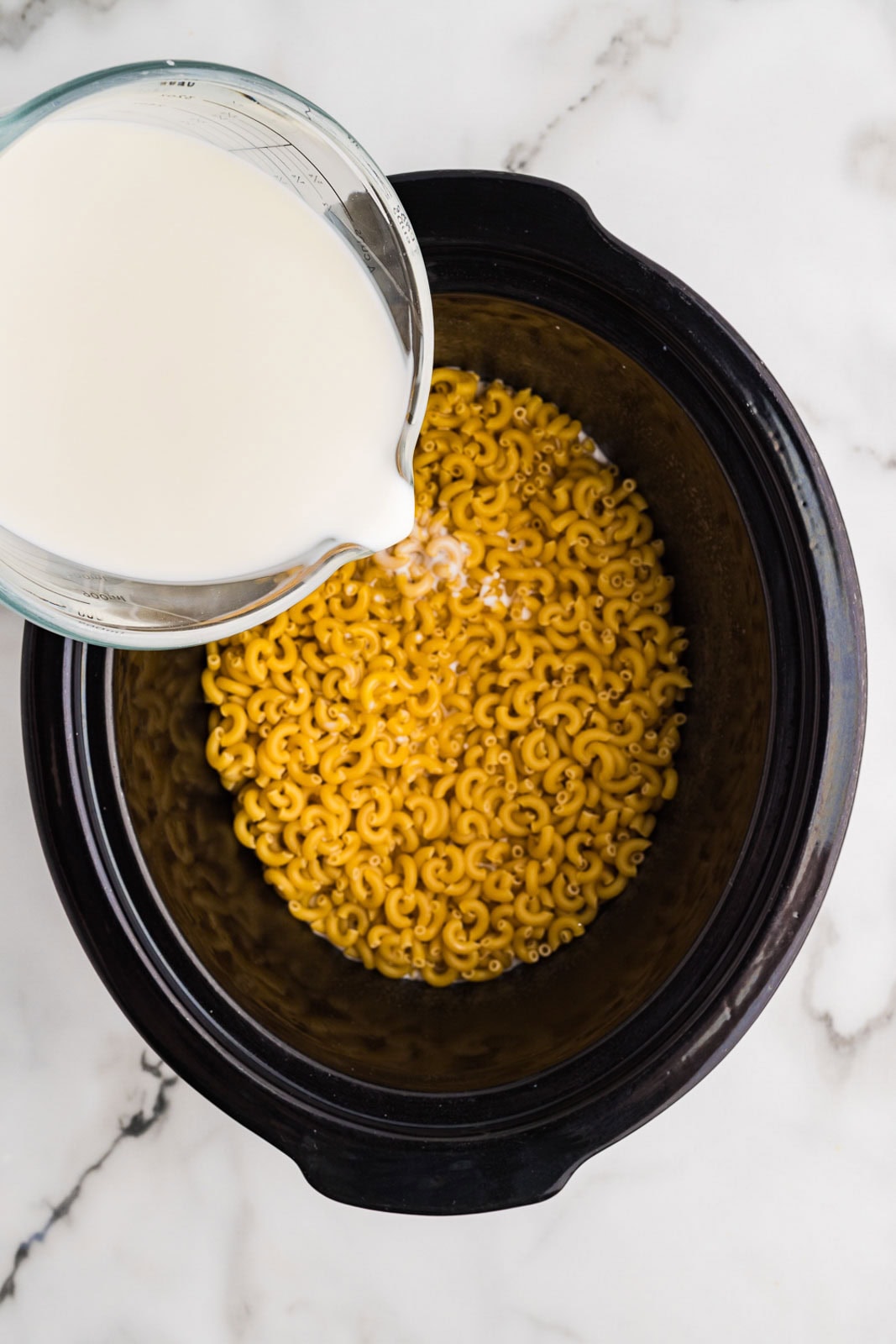 overhead view of black slow cooker with uncooked macaroni and glass measuring cup of milk being poured into slow cooker