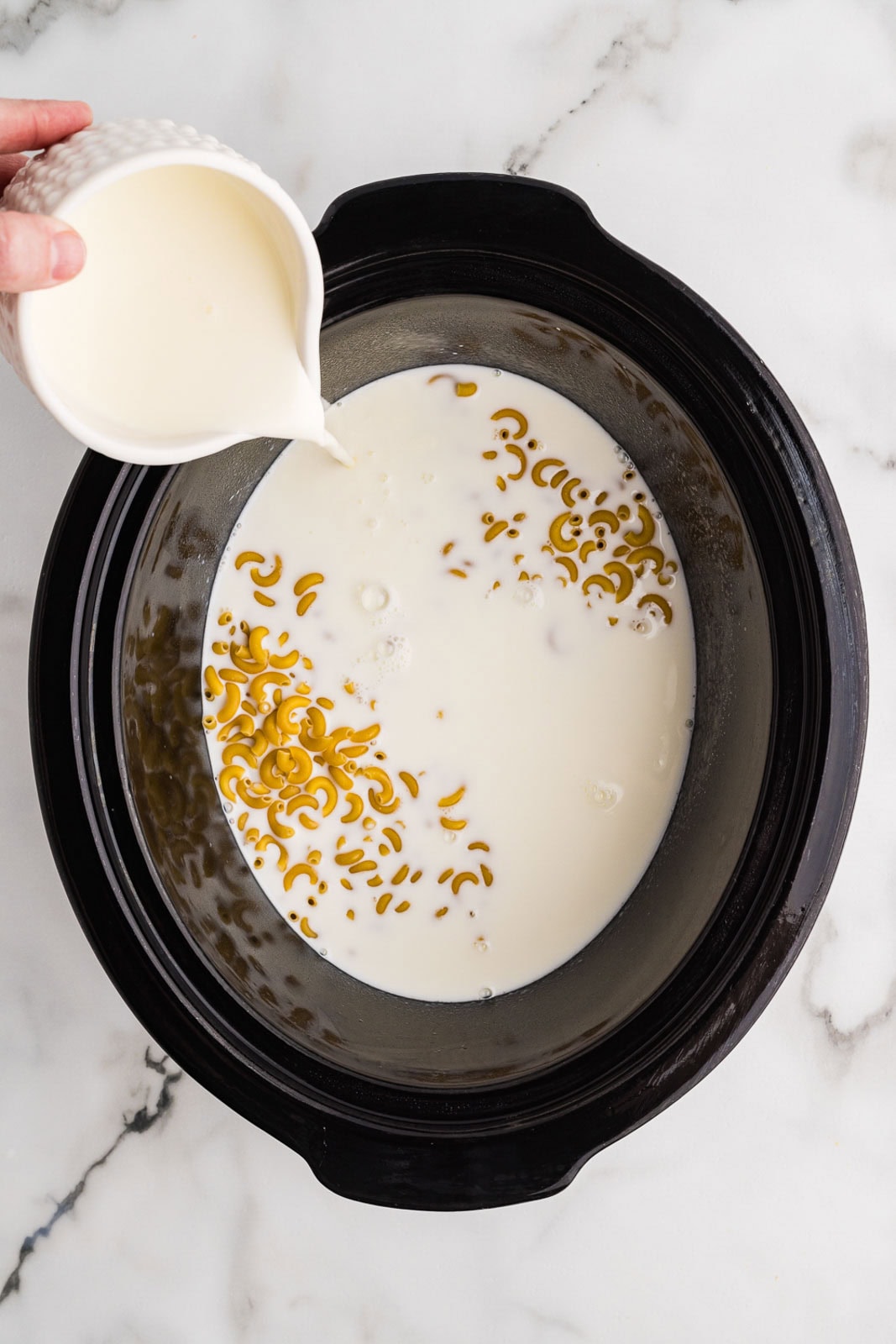 overhead view of black slow cooker with uncooked macaroni, milk and container of cream being poured into mixture