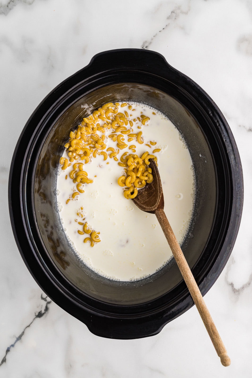 overhead view of black slow cooker with uncooked macaroni with cream and milk being stored with a wooden spoon
