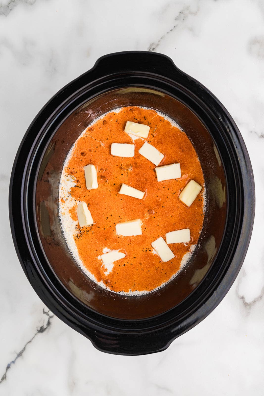 overhead view of black slow cooker with uncooked macaroni with seasonings and pats of butter on top