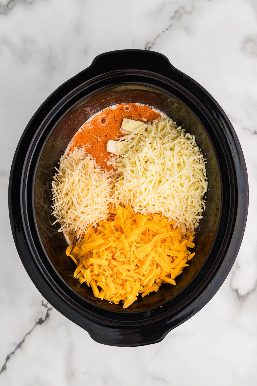 overhead view of black slow cooker with uncooked macaroni with seasonings, butter and all cheeses on top