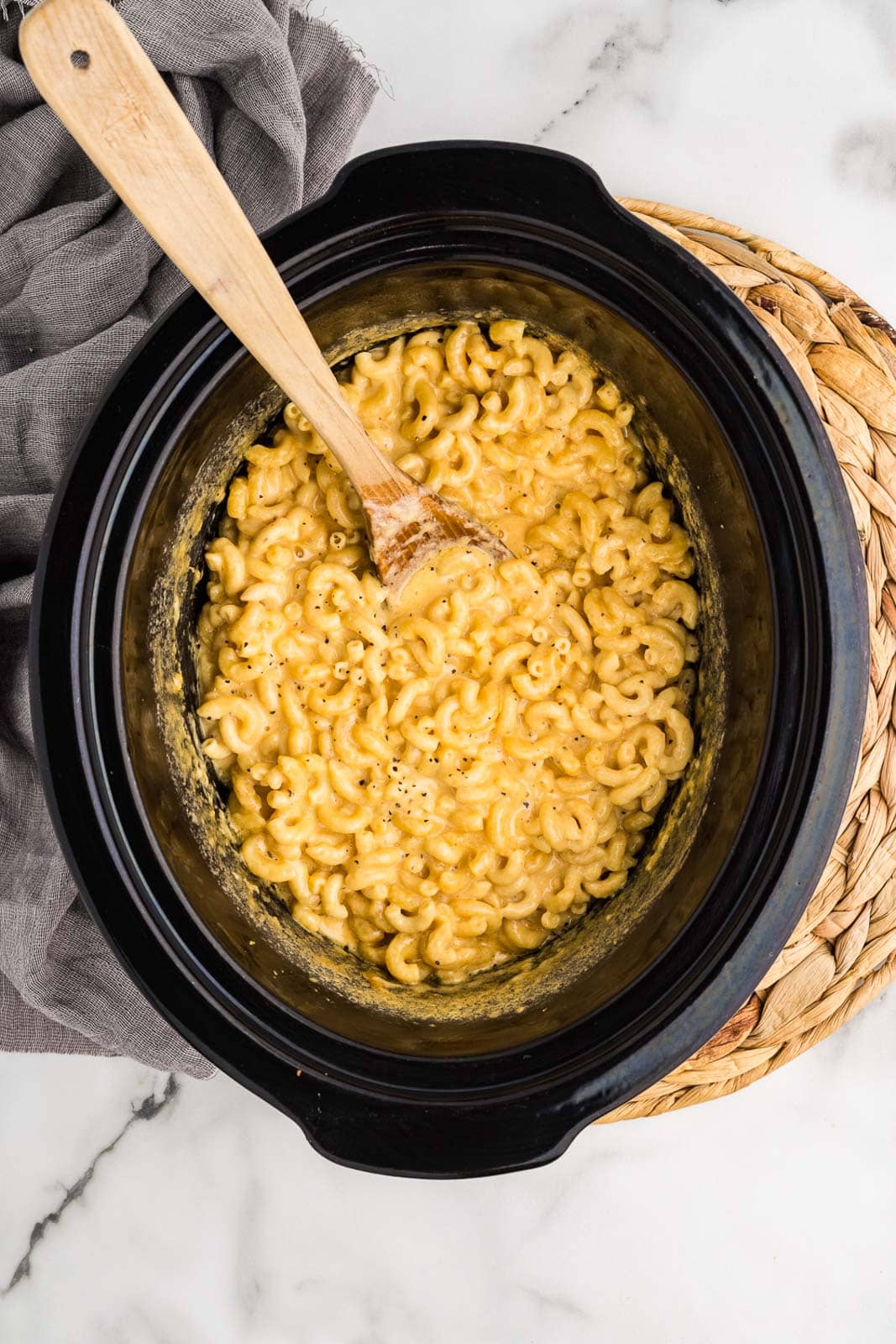overhead view of black slow cooker on a placemat, with cooked Mac and cheese with wooden spoon inserted