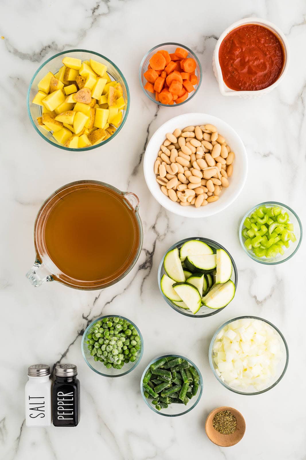 vegetable soup ingredients to include bowls of diced onion, sliced carrots, sliced celery, diced yukon potatoes, sliced zucchini, marinara sauce, white kidney beans, green beans, peas, measuring glass of vegetable broth, wooden bowl of italian seasoning, and salt and pepper shakers