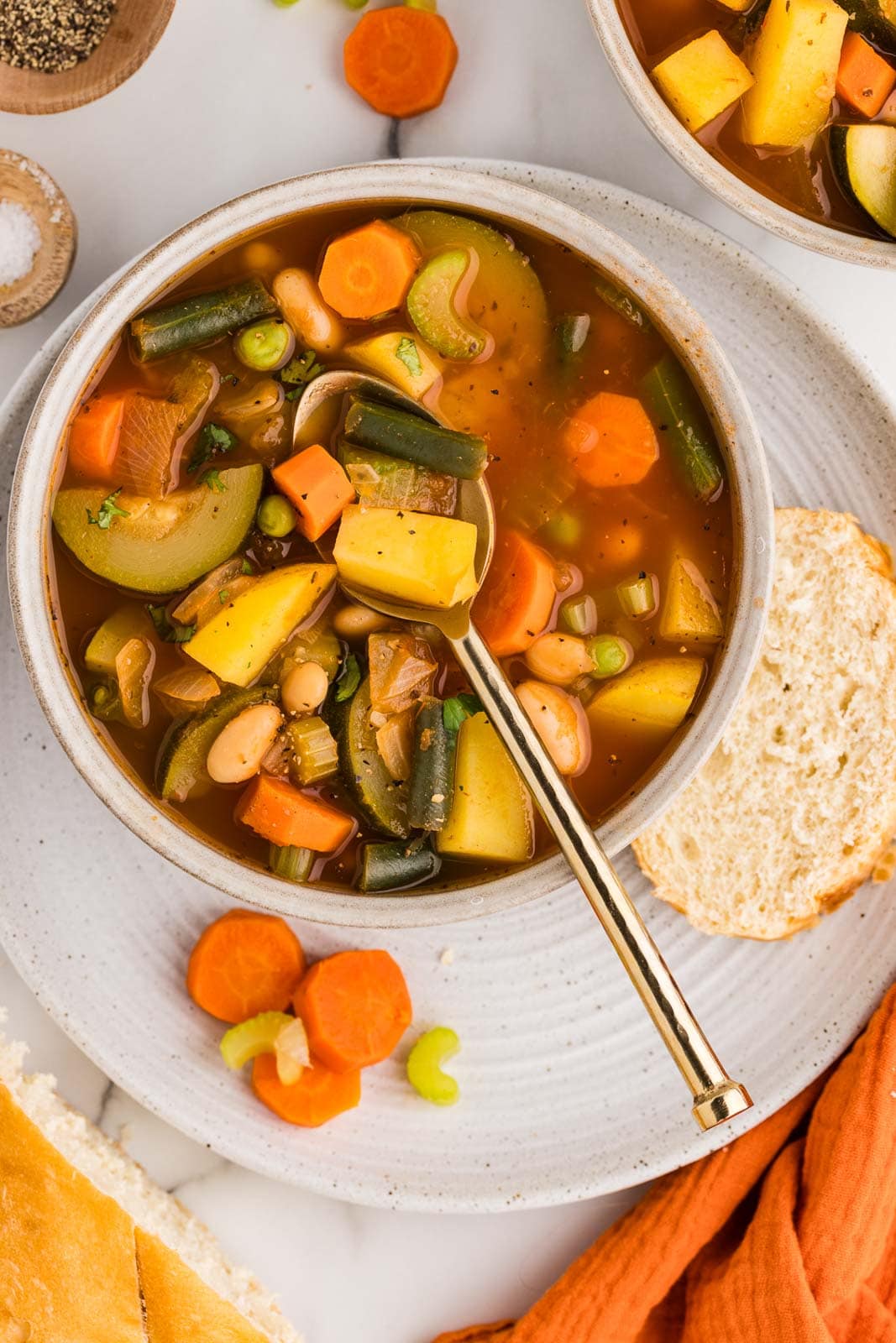 overhead view of bowl of vegetable soup with gold spoon inserted sitting on a plate with a slice of bread, surrounded by another bowl of soup, wooden bowls of seasoning and salt, and orange tea towel