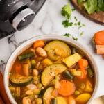 overhead view of bowl of vegetable soup, surrounded by gold spoon, wooden cutting board with parsley, and black slow cooker in background