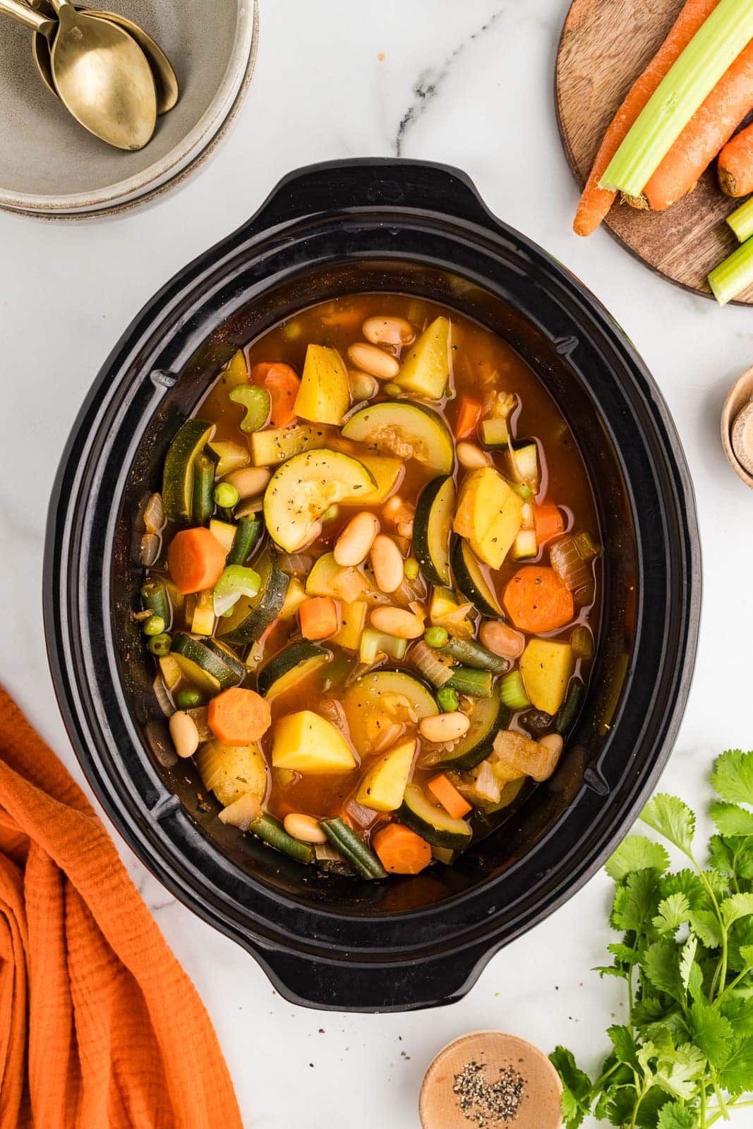 overhead view of black slow cooker with cooked vegetable soup surrounded by wooden cutting board with carrots and celery, bowls and spoons, stalk of parsley, and wooden bowl of seasoning