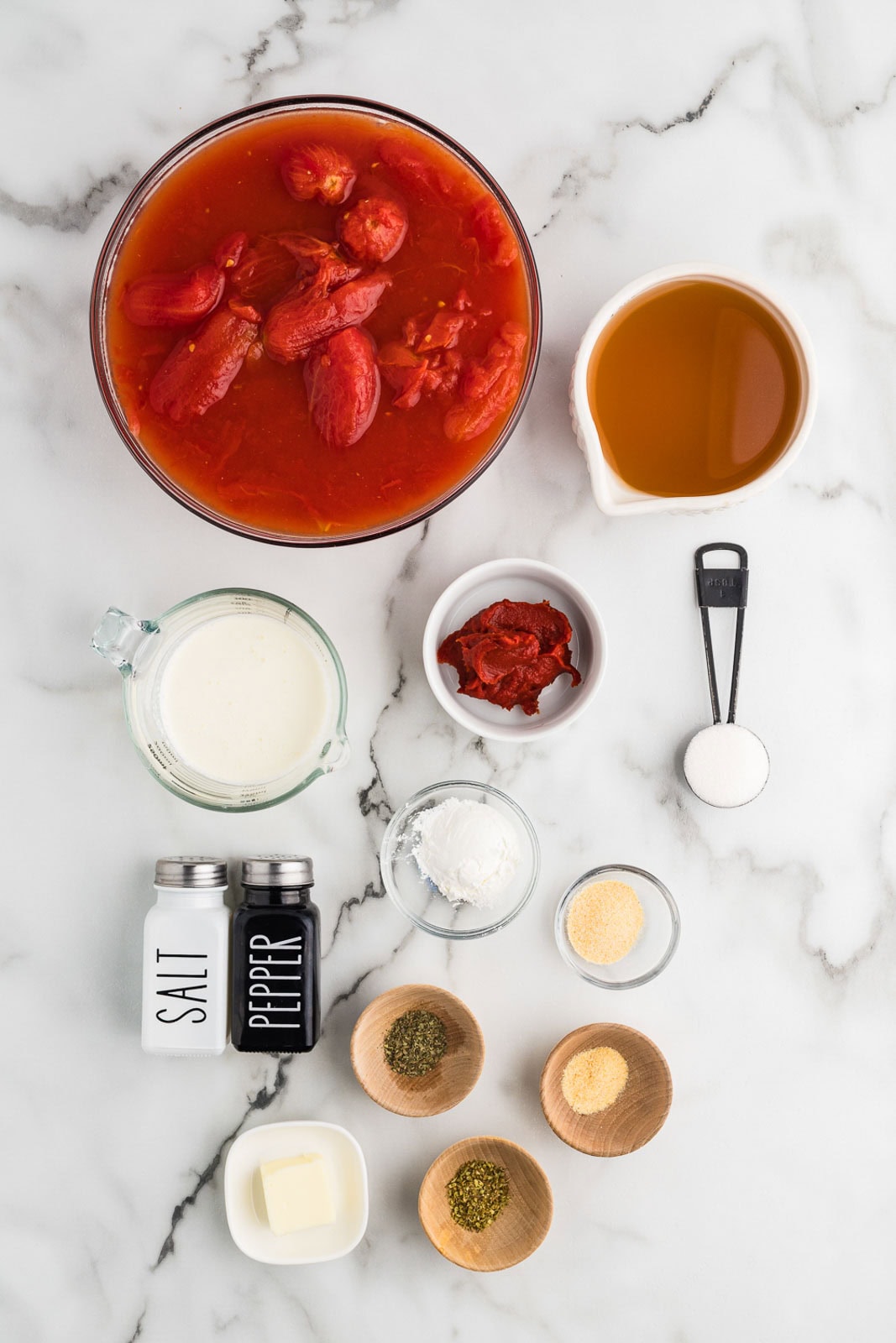 ingredients to include large glass bowl of whole peeled tomatoes surrounded by bowls of tomato paste, vegetable broth, onion and garlic powders, dried basil, dried oregano, heavy cream, butter, tapioca flour, sugar and salt and pepper shakers
