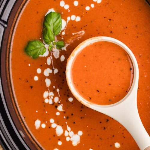 overhead view of black slow Cooker with white ladle inserted into tomato soup with fresh basil on top