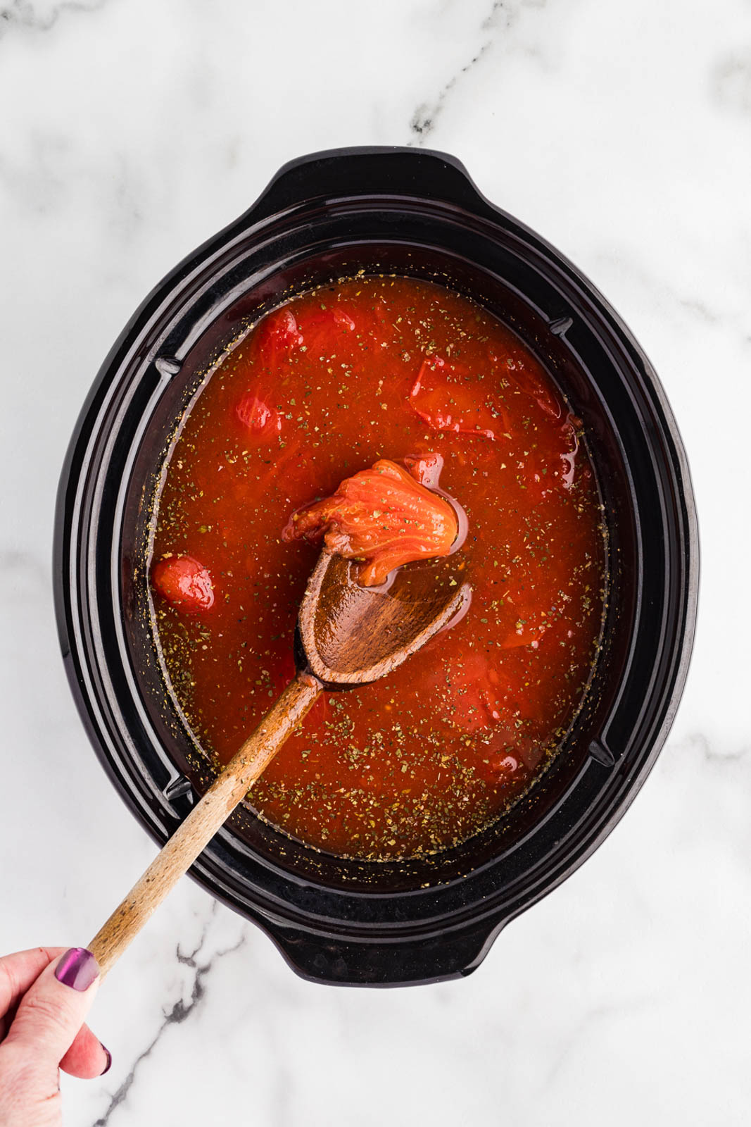 overhead view of black slow Cooker containing all ingredients except for cream, buttery, slurry and sugar, with wooden spoon breaking up the tomatoes