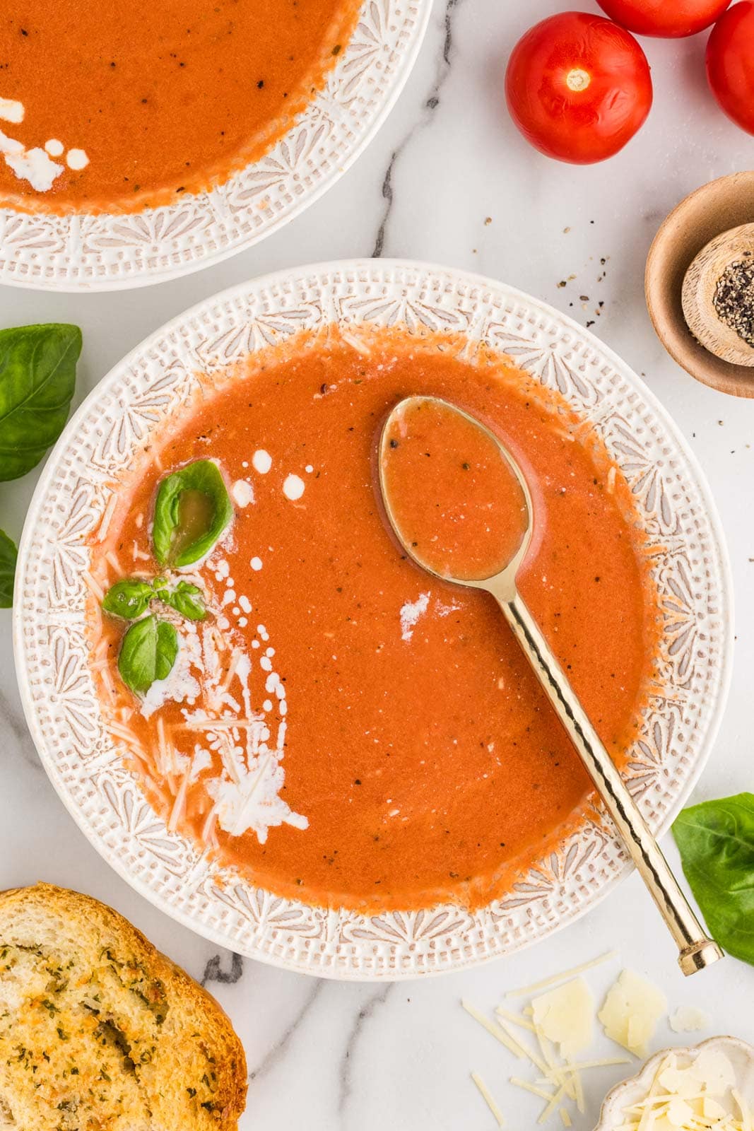 overhead view of white bowl of soup with spoon inserted, with another bowl of soup, crusty bread, and tomatoes on side