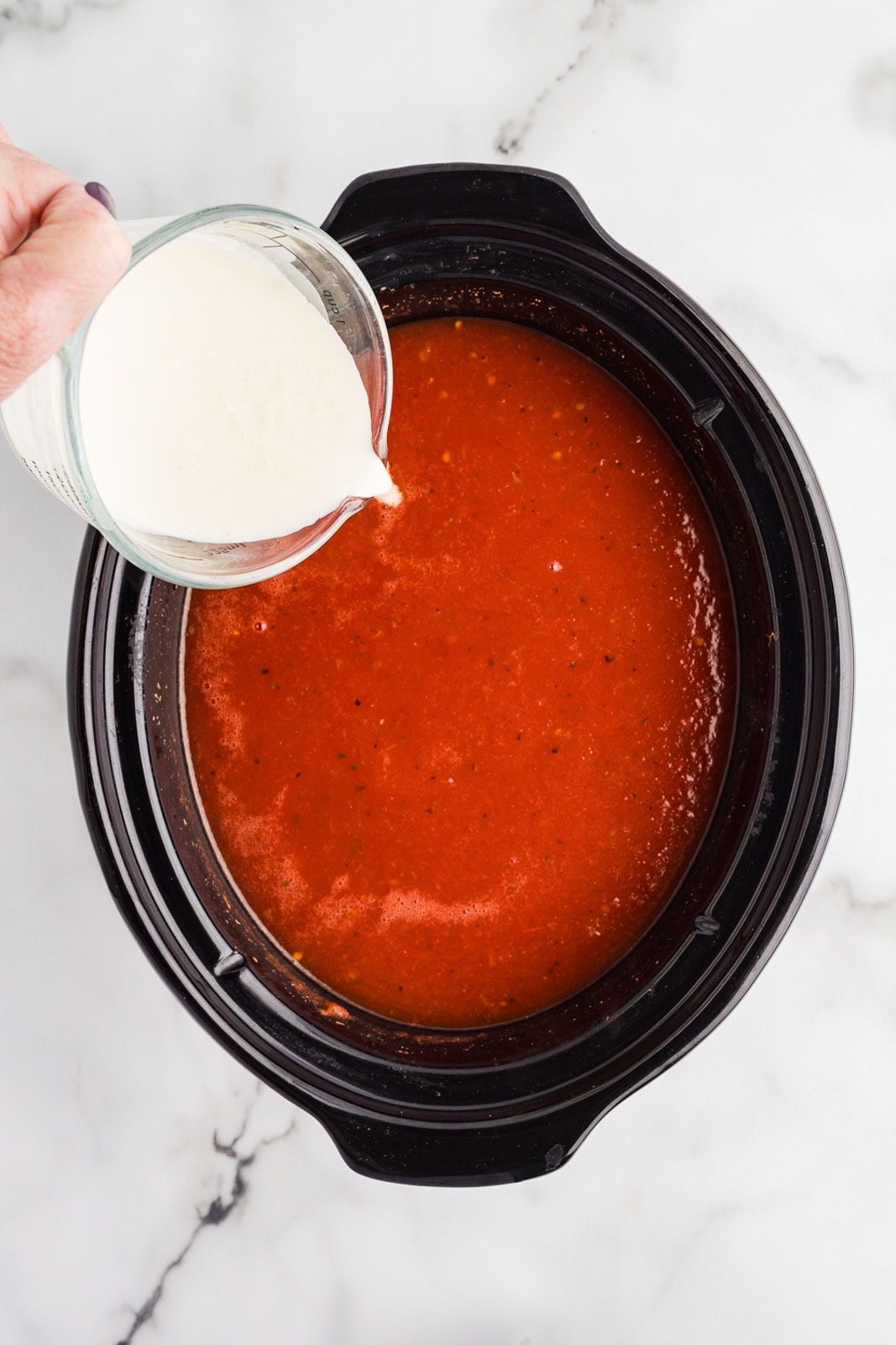 overhead view of black slow Cooker of cooked and blended mixture with hand holding heavy cream being poured into mixture