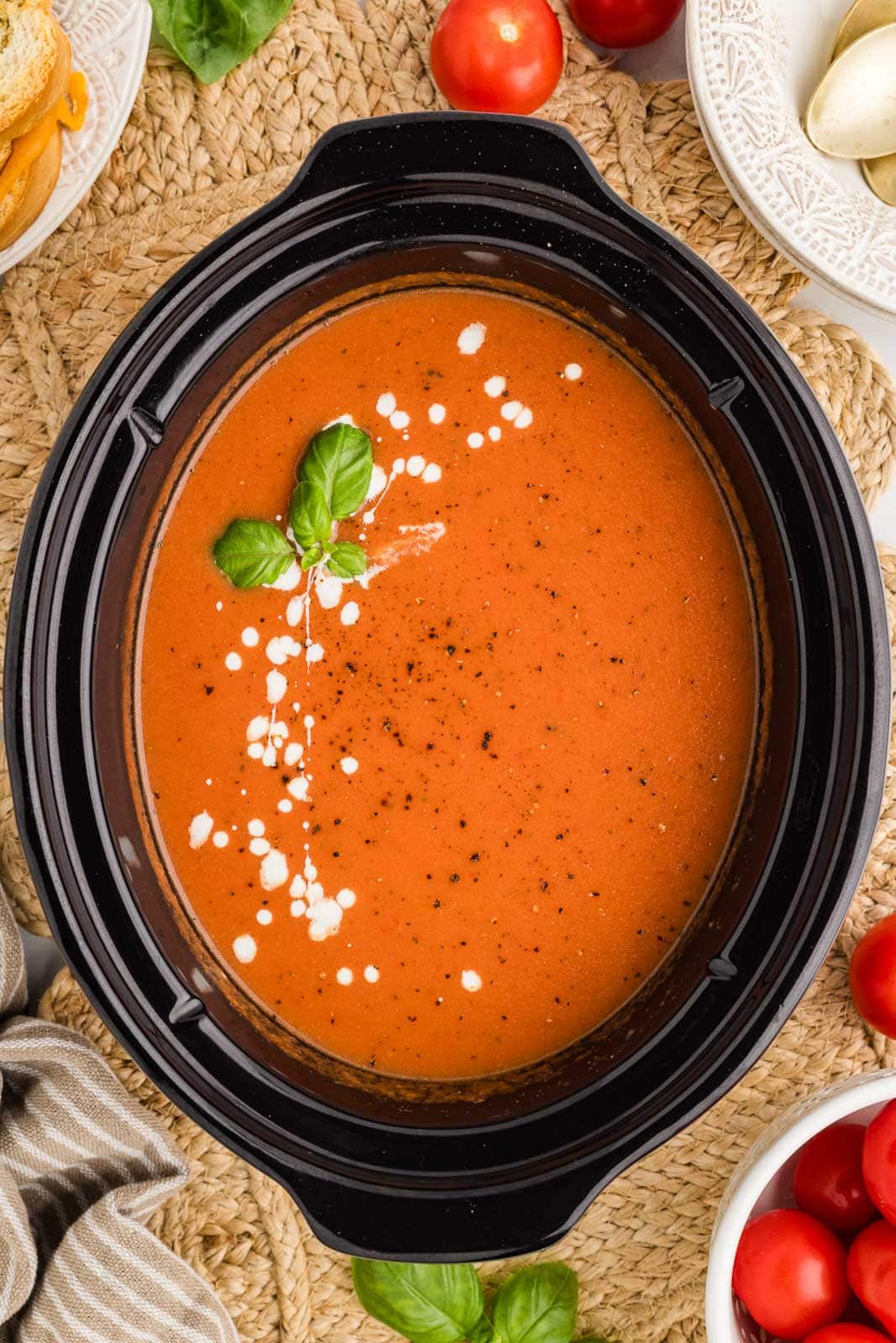 overhead view of black slow Cooker containing slow cooked tomato soup with fresh basil, on top of place mat surrounded by bowls of tomatoes, basil, bread