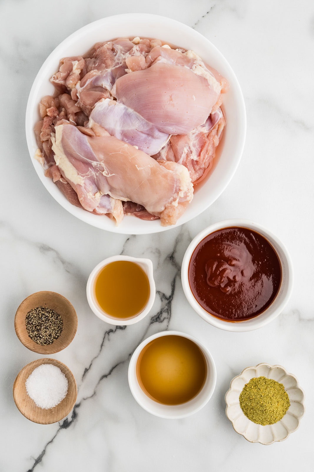 ingredients to include large white bowl of chicken thighs surrounded by bowls of herb and garlic seasoning, salt, pepper, chicken broth, bbq sauce and apple cider vinegar