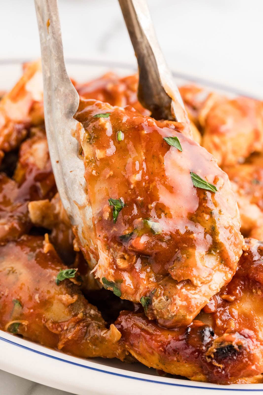 closeup of slow cooked bbq chicken thighs in white serving platter with silver tongs