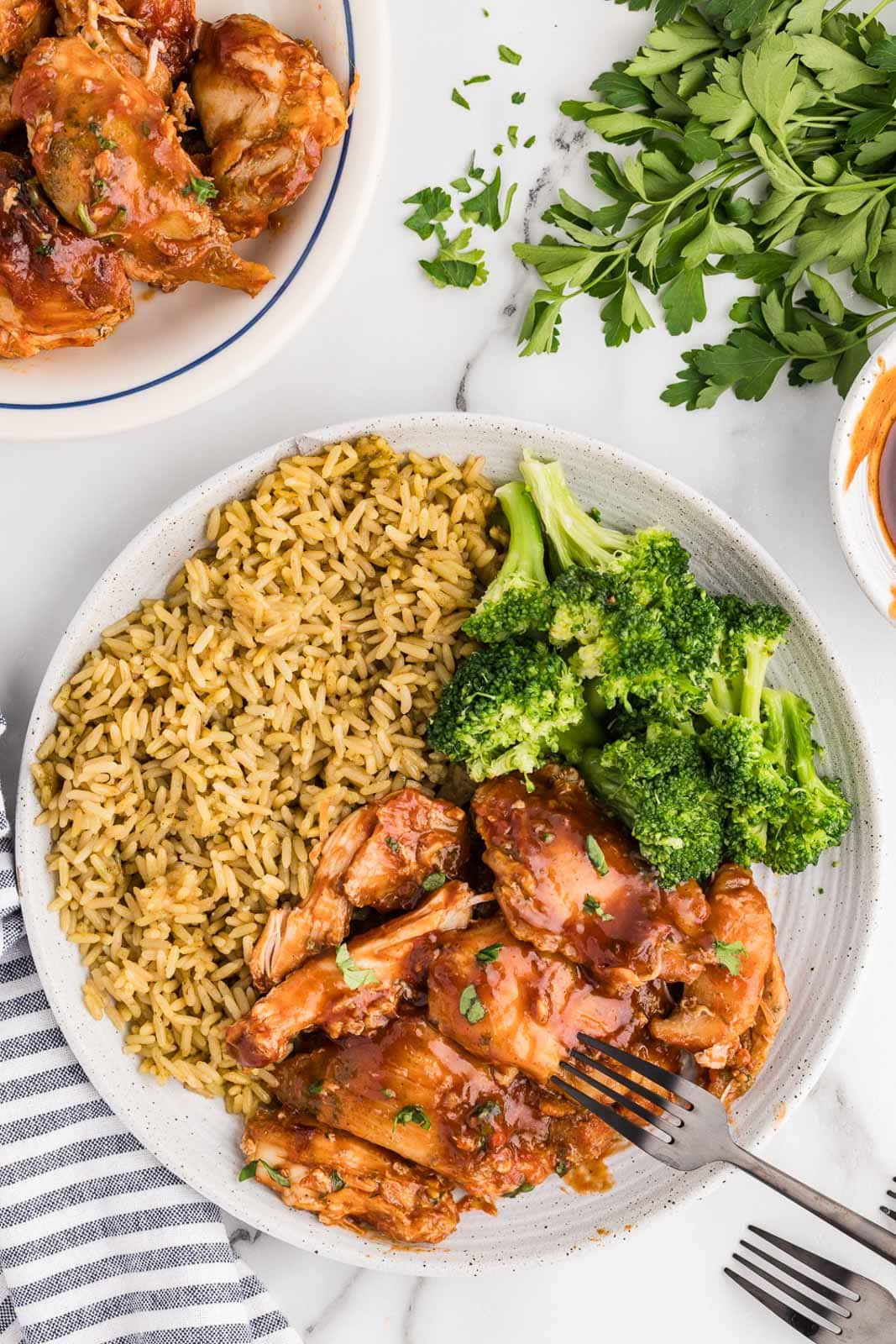 overhead view of dinner plate of bbq chicken thighs, broccoli and rice with bowl of chicken thighs and parsley on side