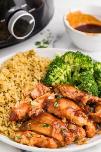 closeup view of white dinner plate of bbq chicken thighs, broccoli and rice with bowl of bbq sauce and black slow Cooker in background