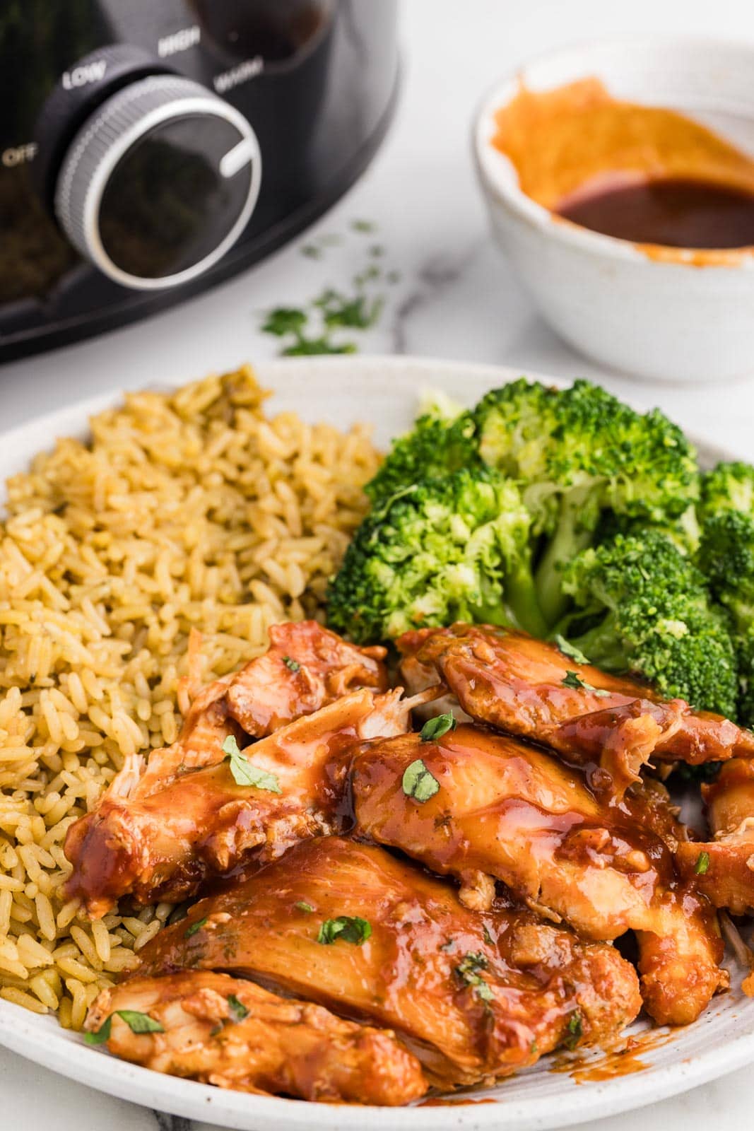 closeup view of white dinner plate of bbq chicken thighs, broccoli and rice with bowl of bbq sauce and black slow Cooker in background