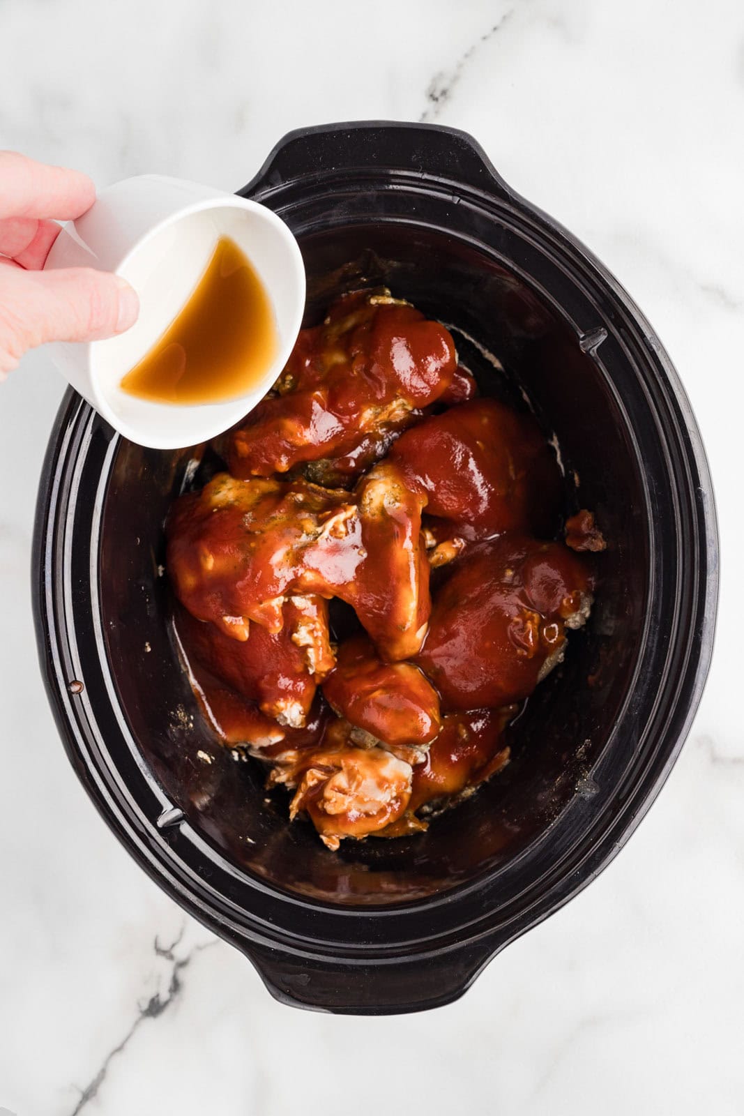 overhead view of black slow Cooker with slow cooked chicken thighs with bbq sauce and hand holding small white bowl of apple cider vinegar being poured over thighs