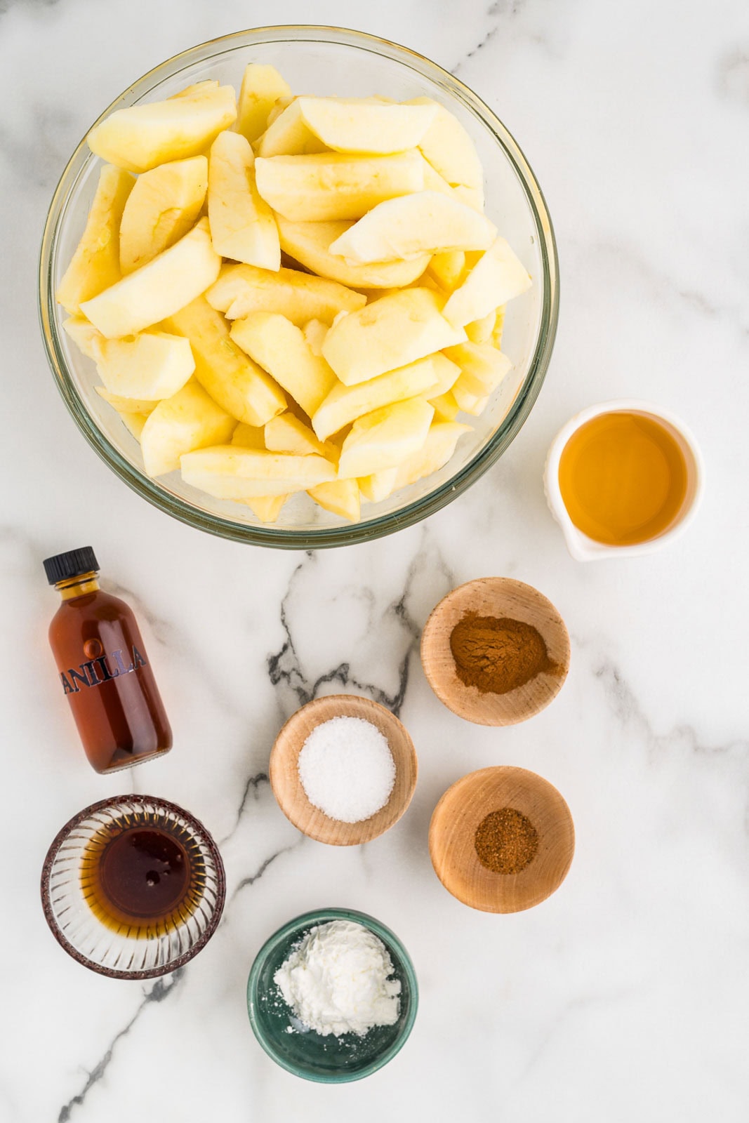 ingredients to include large glass bowl of peeled and sliced apples, with small bowls of apple juice, maple syrup, ground cinnamon, ground nutmeg, salt, tapioca and a small container of vanilla