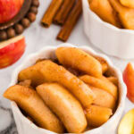 closeup view of white ramekin of slow cooked apples with another ramekin in background, with apple slices, gold spoon, cinnamon sticks, whole apple surrounding it