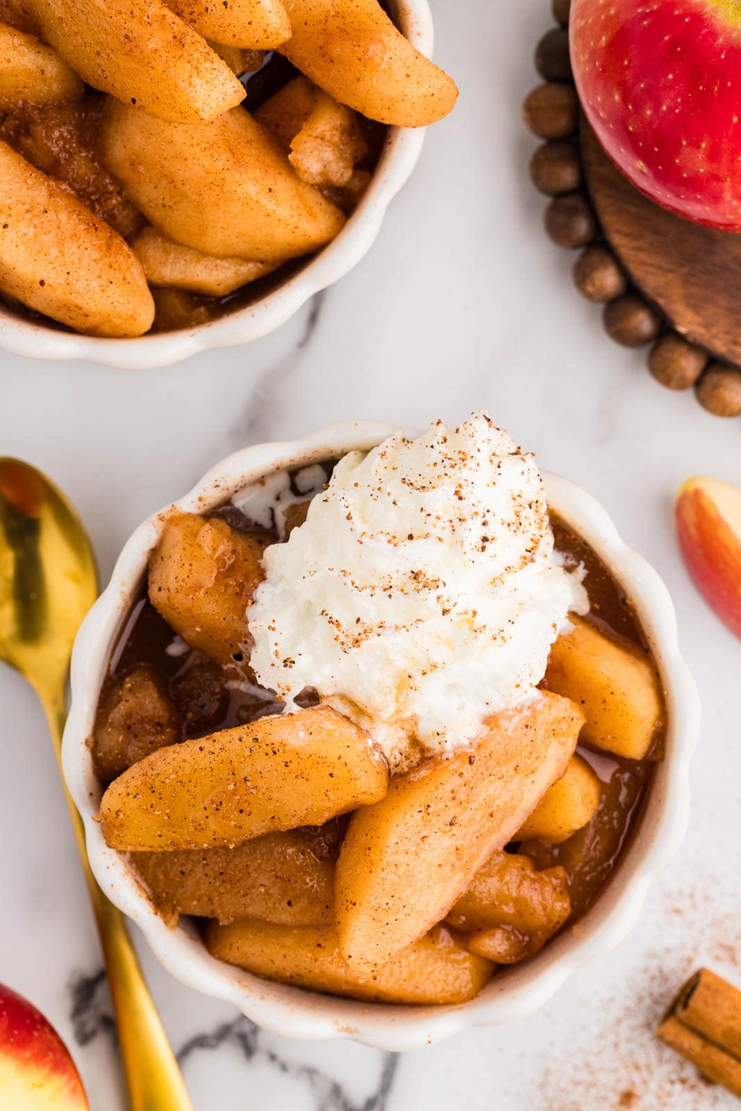overhead view of white ramekin of cooked apples topped with vanilla ice cream with another ramekin in background, surrounded by gold spoon, whole apple, and cinnamon sticks