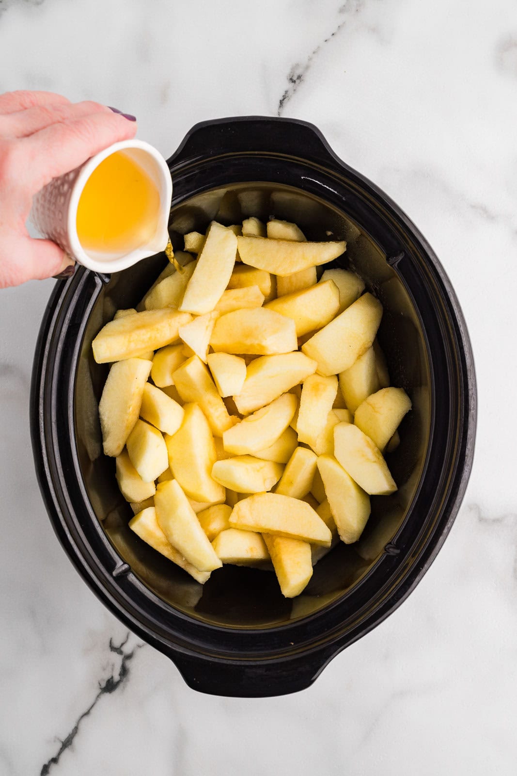 overhead view of black slow Cooker with sliced apples with hand pouring apple juice onto apples