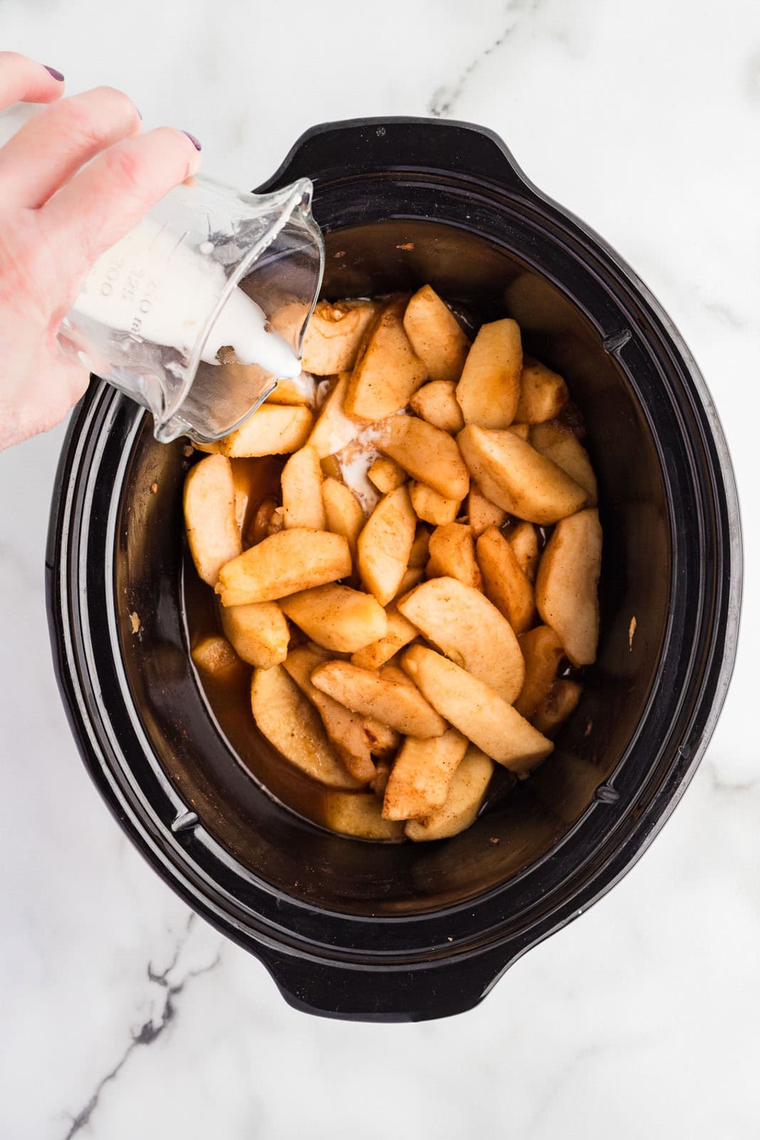 overhead view of black slow Cooker with cooked apples with hand pouring tapioca and vanilla slurry over apples