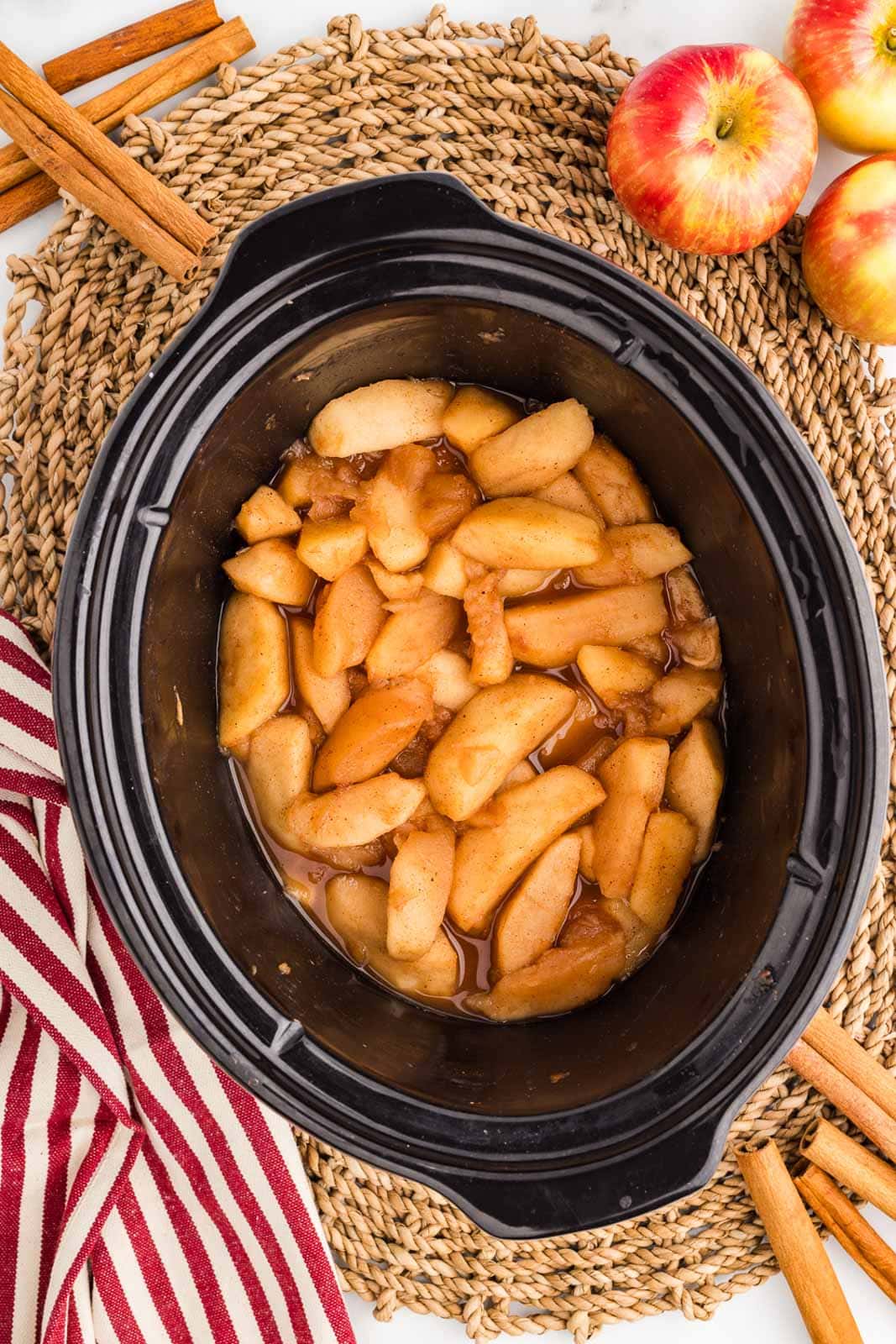 overhead view of slow cooked apples on a woven place mat surrounded by apples, cinnamon sticks and striped dish towel