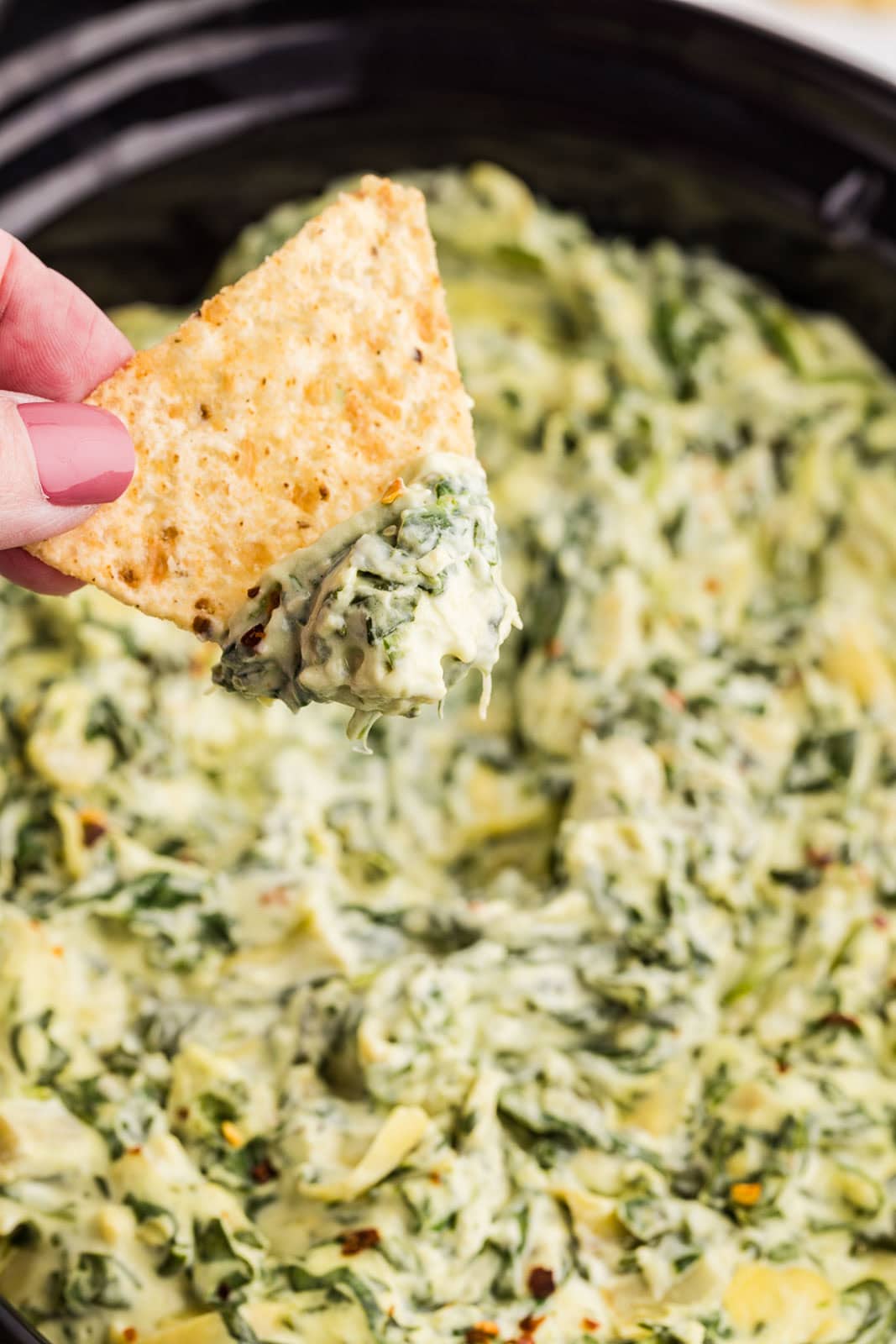 closeup of hand holding chip and dip, with black slow cooker of dip in background