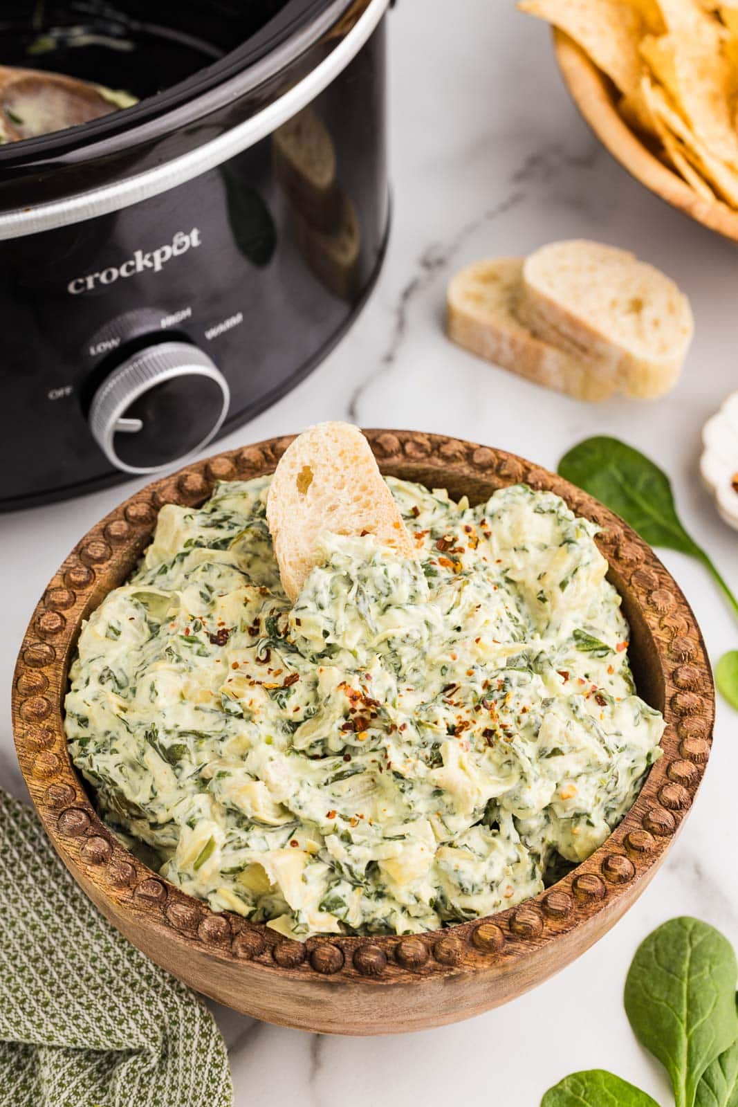 Overhead view of serving bowl of dip and a slice of bread, with black slow cooker, bread slices and bowl of tortilla chips in background