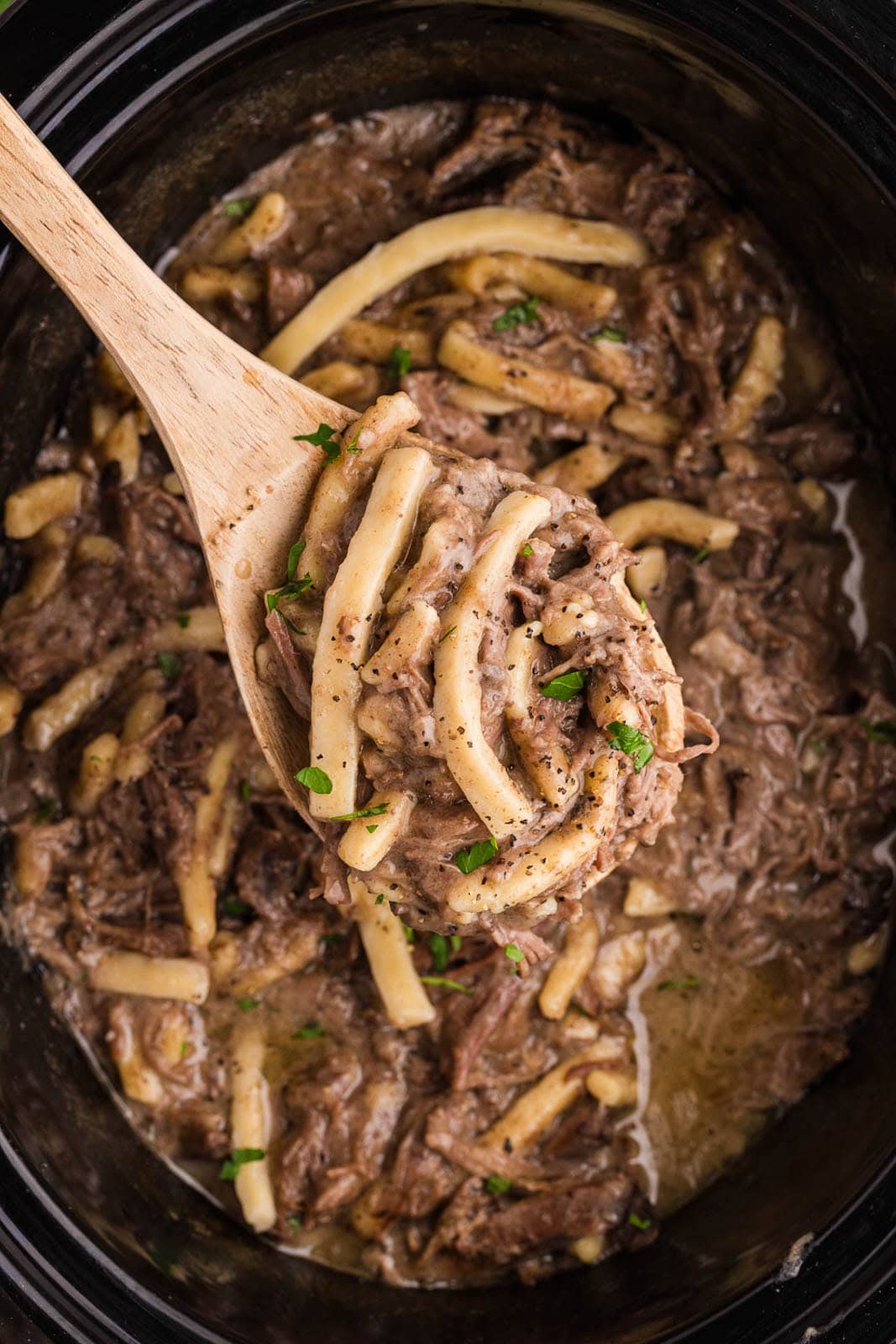 closeup view of wooden spoon of beef and noodles with black slow cooker of beef and noodles in background