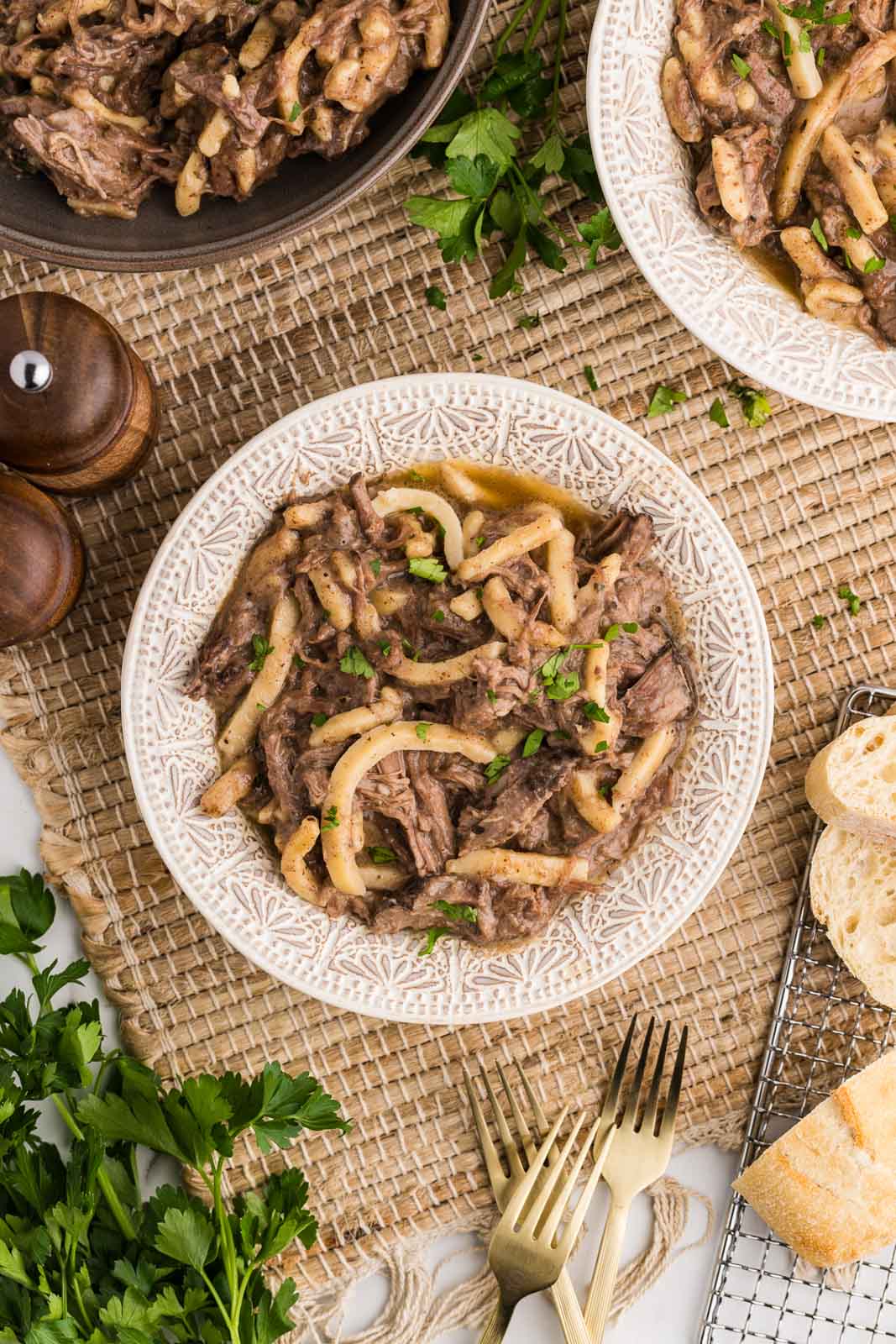 overhead view of two dinner plates of beef and noodles surrounded by larger bowl of beef and noodles, salt and pepper shakers, forks and sliced bread on side