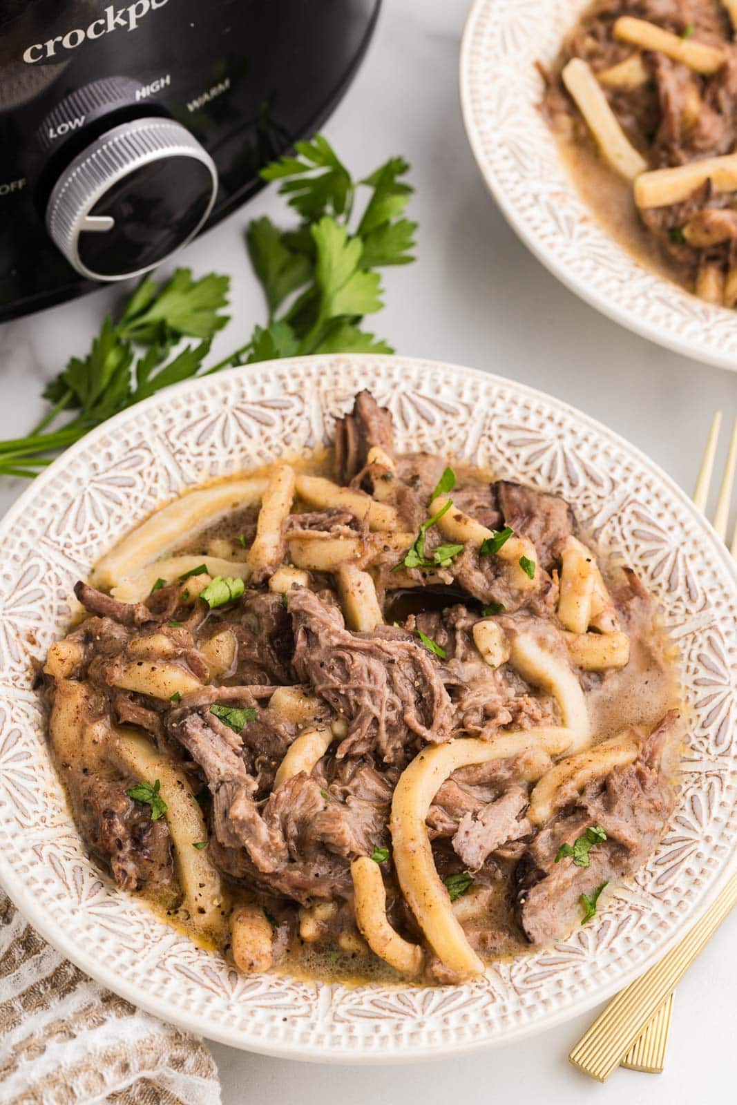 dinner plate of beef and noodles with another in background along with black slow cooker