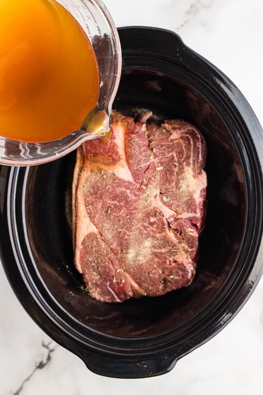 overhead view of seasoned roast in black slow cooker with beef broth being poured into slow cooker