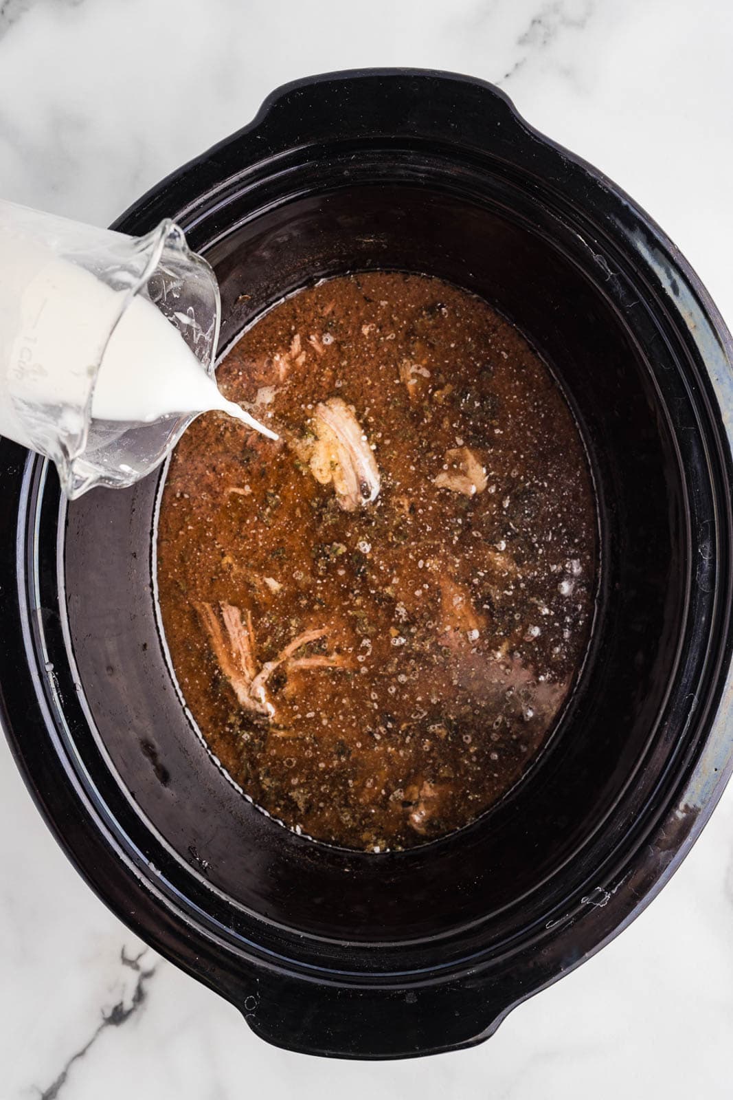 overhead view of black slow cooker of roast with container of cornstarch slurry being poured into slow cooker