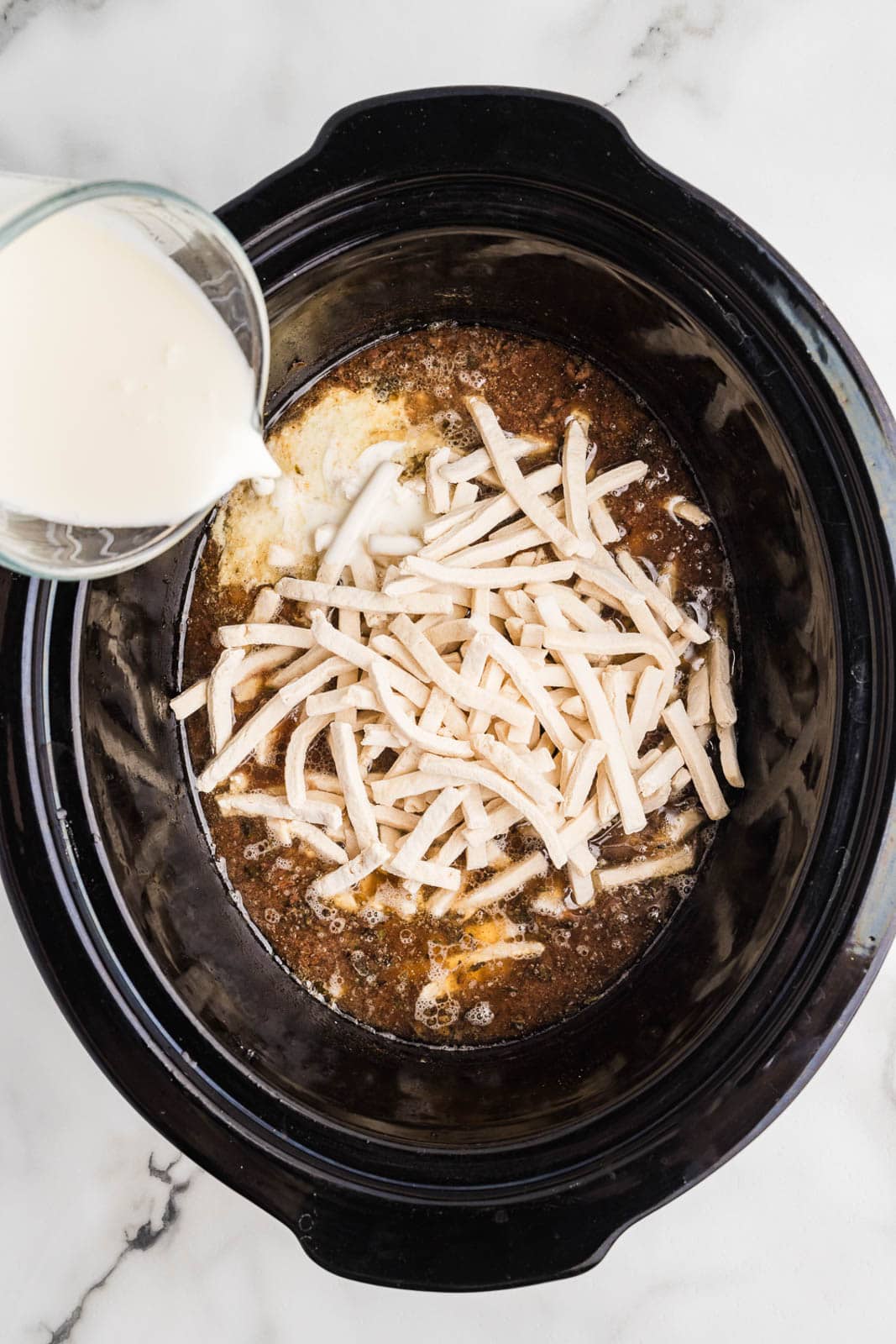 overhead view of black slow cooker with noodles and roast with heavy cream being poured into slow cooker
