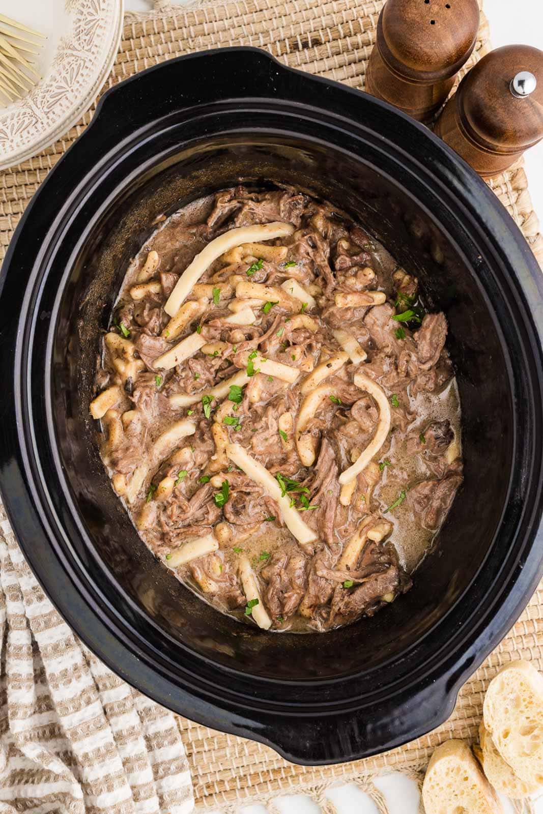 overhead view of cooked beef and noodles with parsley sprinkled on top with salt and pepper shakers and slices of bread on side