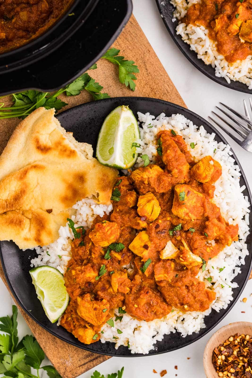 overhead view of black dinner plate of Tikka masala on bed of white rice, pita and lime wedges on a wooden cutting board, with black slow cooker and another dinner plate on side