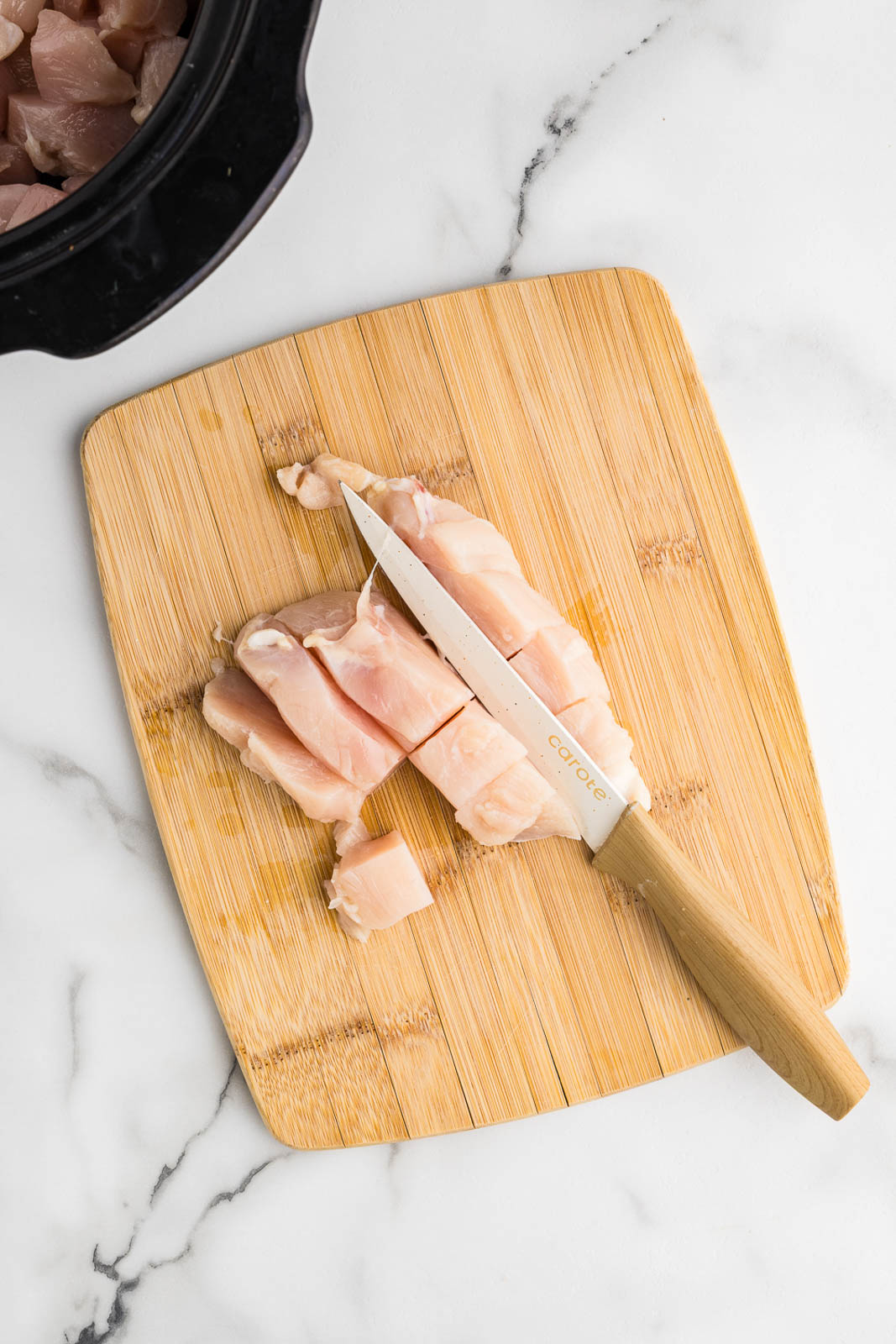 wooden cutting board with chicken breast being cut into cubes with knife, with black slow cooker in background