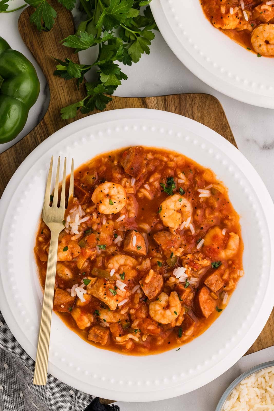 overhead view of white dinner bowl with jambalaya with fork surrounded by green pepper, parsley and another dinner plate