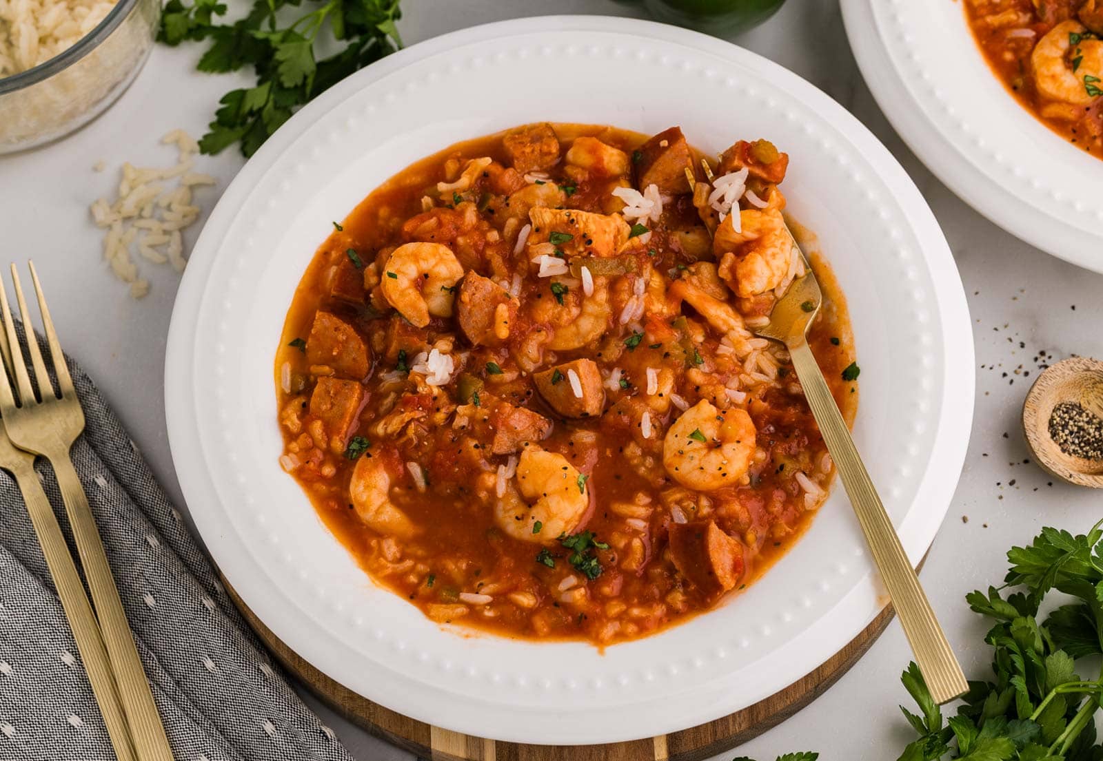 closeup overhead view of white dinner bowl of jambalaya with fork inserted surrounded by 2 forks, another dinner bowl of jambalaya, parsley, bowl of pepper, and bowl of cooked rice
