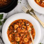 overhead view of 2 dinner bowls of jambalaya with forks with black slow cooker of jambalaya in background