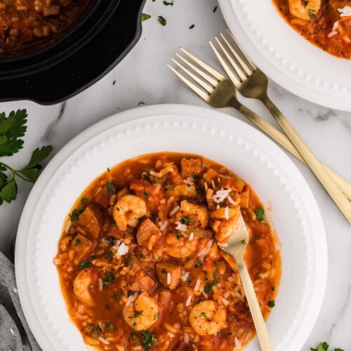 overhead view of 2 dinner bowls of jambalaya with forks with black slow cooker of jambalaya in background