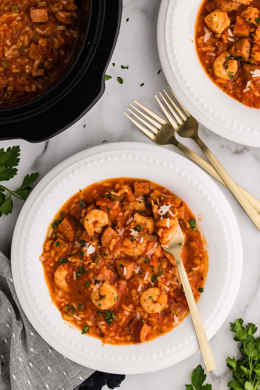 overhead view of 2 dinner bowls of jambalaya with forks with black slow cooker of jambalaya in background