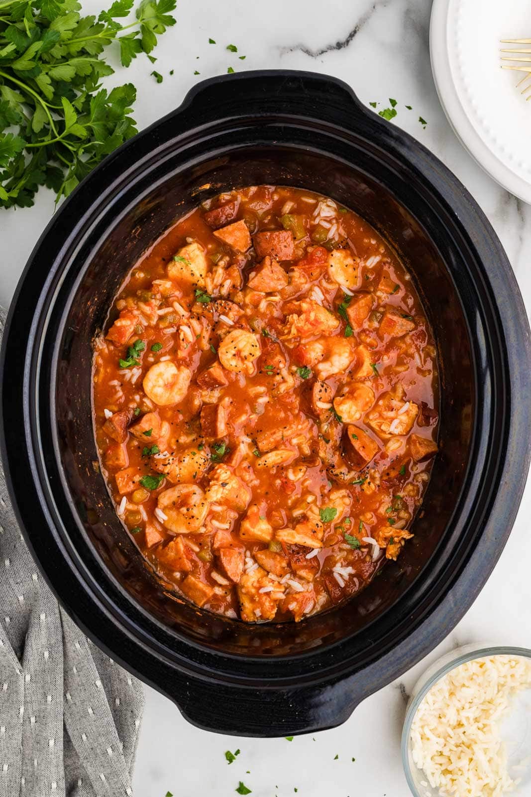 black slow cooker with slow cooked jambalaya surrounded by parsley, hand towel, white dinner plate and small bowl of cooked rice